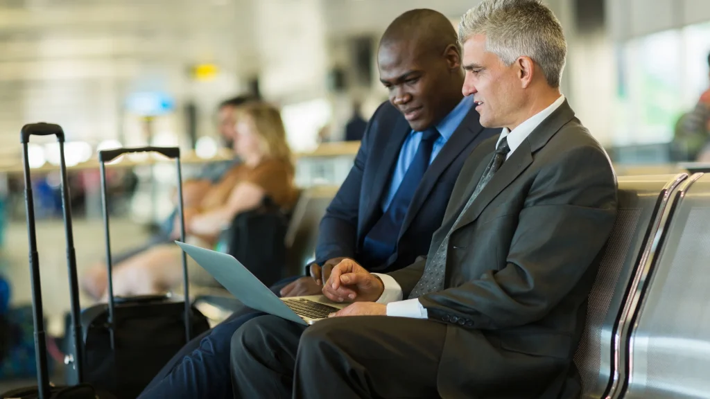 Two diverse businessmen in suits collaborating and working on a laptop in an airport lounge, demonstrating best business travel practices for remaining productive on the road.