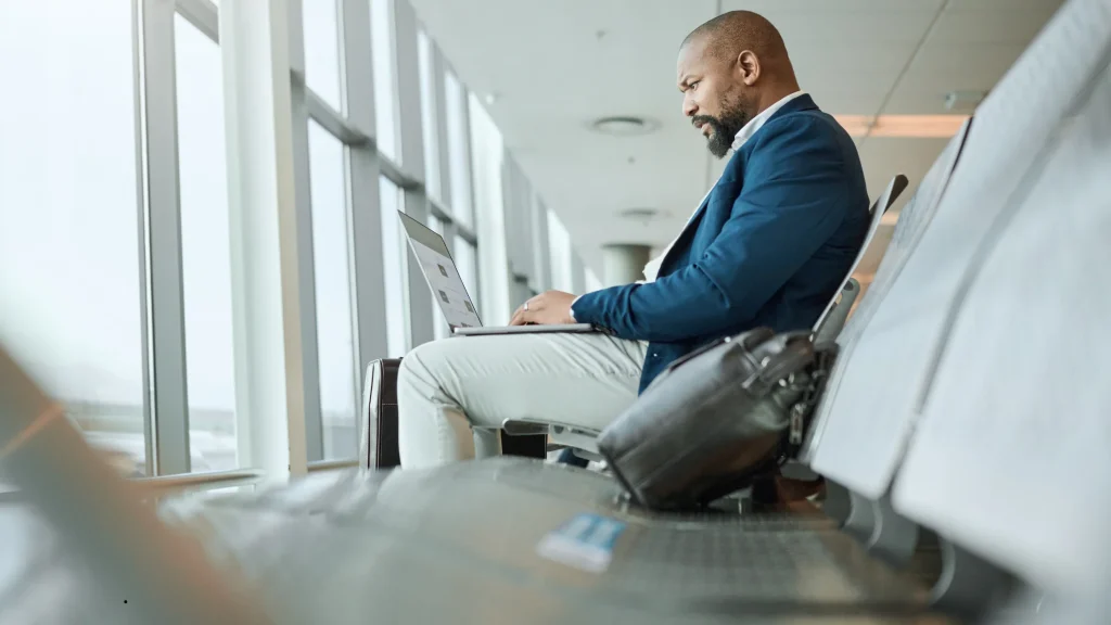 A professional businessman in a blue blazer working on his laptop at an airport terminal departure gate with his luggage nearby, managing a corporate travel booking before his flight.