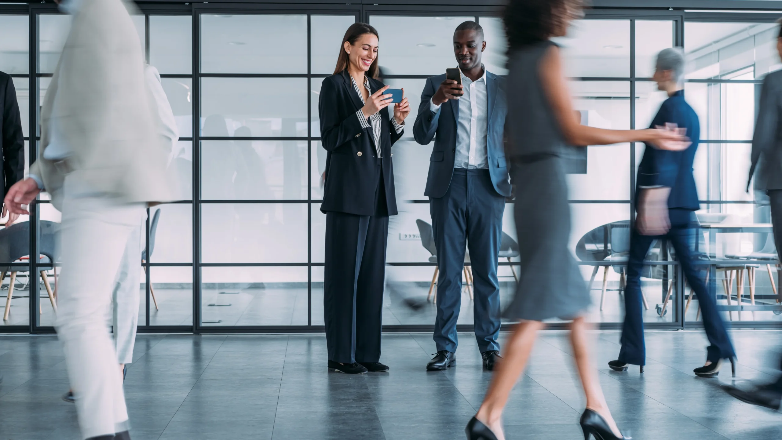 A businessman and businesswoman standing in a busy, modern office lobby looking at their smartphones, managing a corporate travel booking or flight itinerary on the go while colleagues walk past with motion blur.