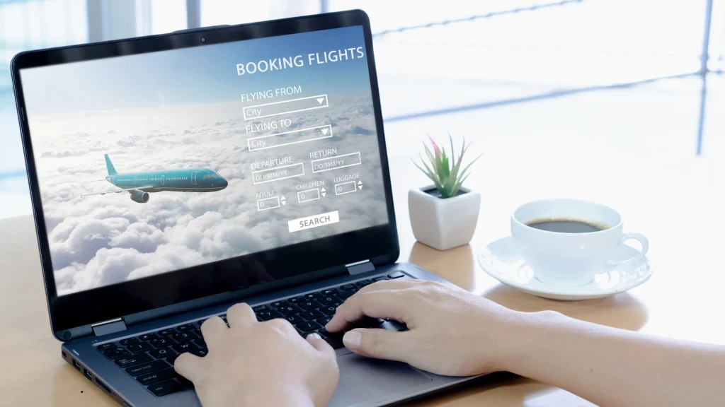 Close-up of hands typing on a laptop displaying a flight search and reservation portal, illustrating how companies manage corporate travel booking from an office desk.