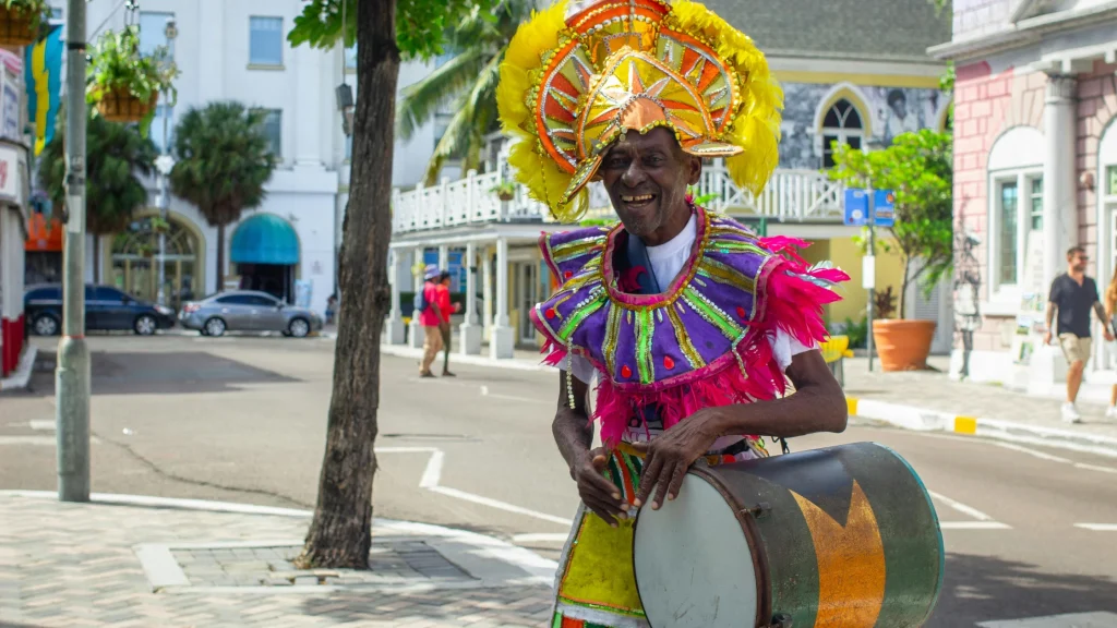 A smiling older Black man in a vibrant, elaborate Junkanoo festival costume stands on a paved city street while playing a traditional drum. He wears a large, ornate yellow and orange feathered headdress and a matching purple and pink layered collar. In the background, colonial-style buildings, tropical trees, passing cars, and a few pedestrians line the sunlit street.